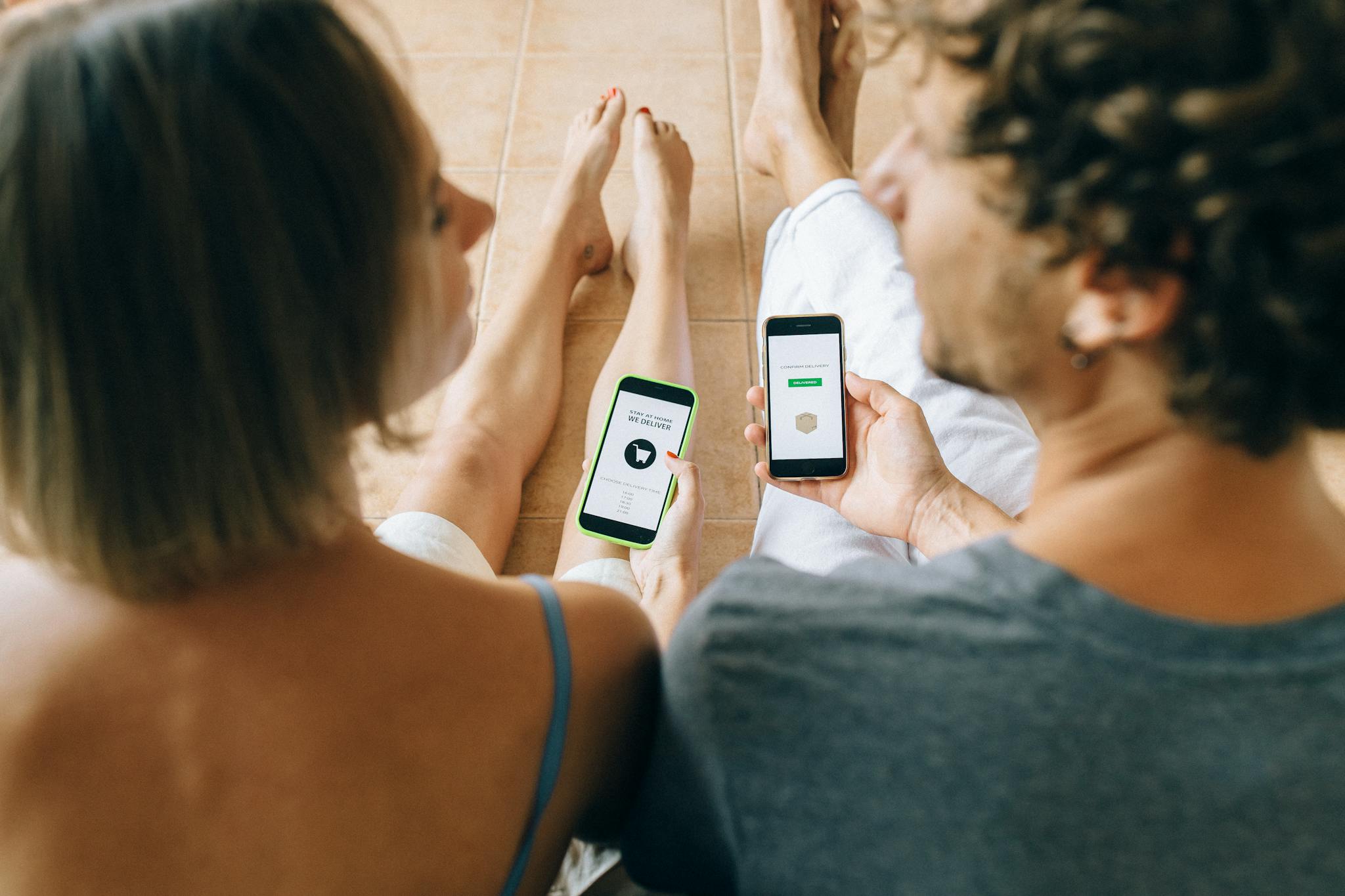 A couple using smartphones for online shopping while relaxing indoors on a tiled floor.