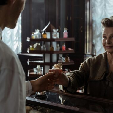 A senior woman receives a perfume bottle from a pharmacist inside a vintage pharmacy.
